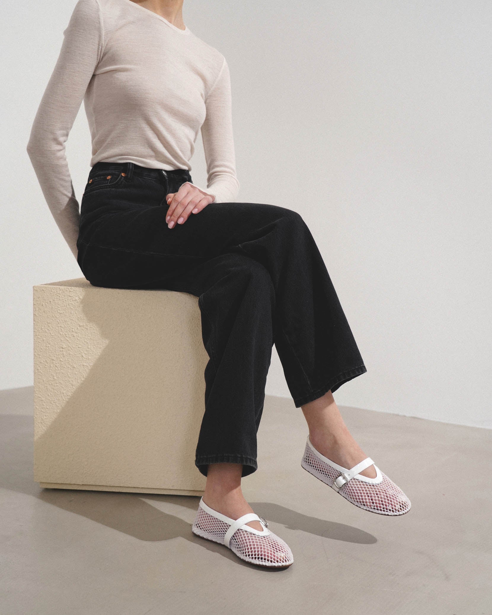 Person wearing black pants and white mesh shoes sitting on a beige cube.
