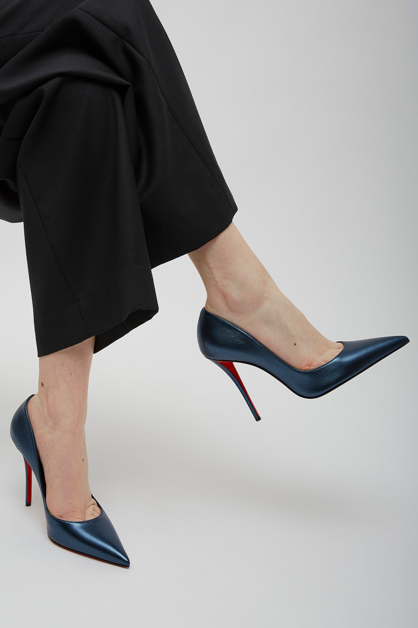 Navy high-heeled shoes with red soles worn by a person in black pants on a light gray background