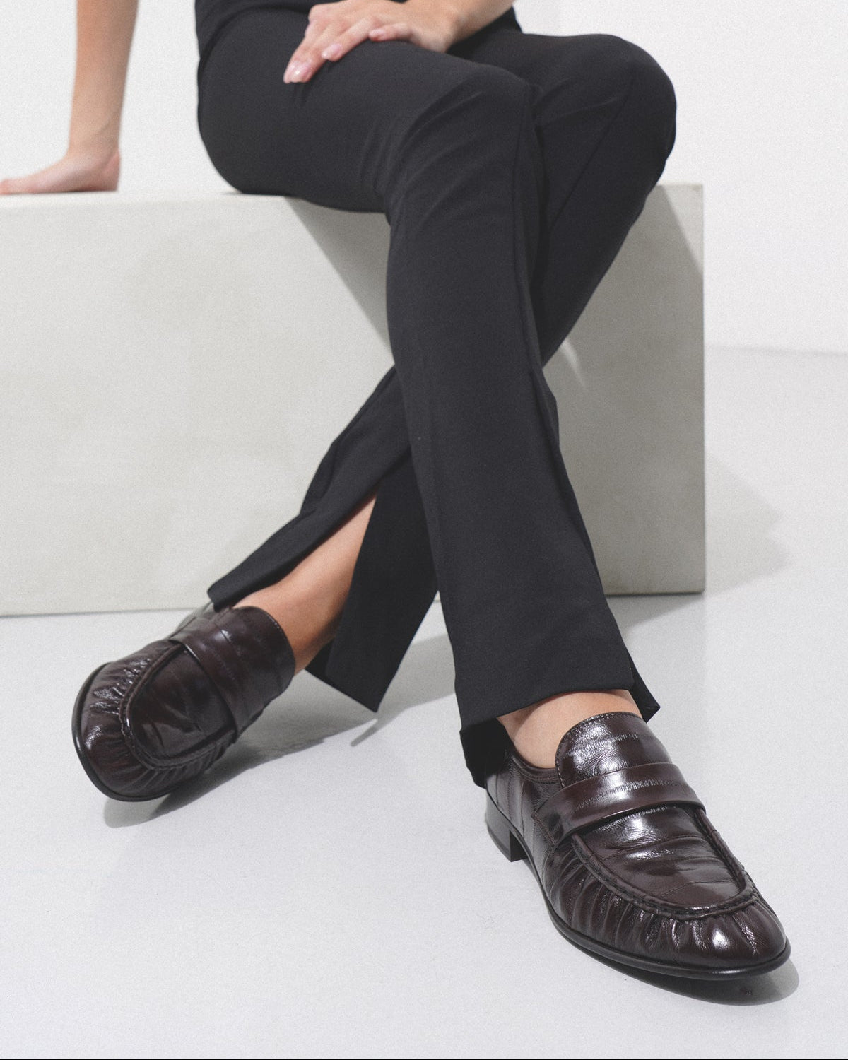 Person wearing black pants and brown shoes sitting on a white surface.