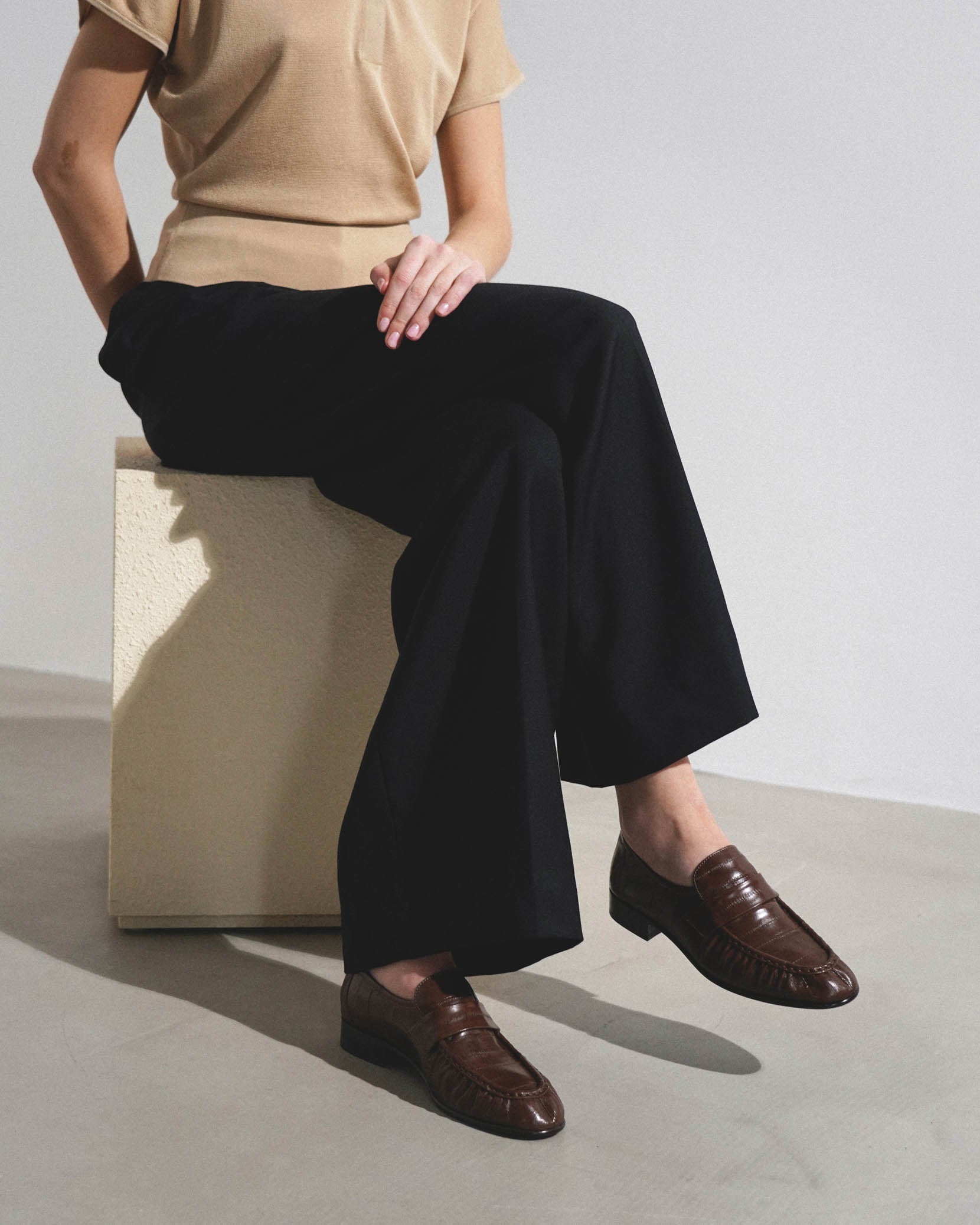 Person wearing a beige top and black wide-leg pants with brown shoes, sitting on a white box.