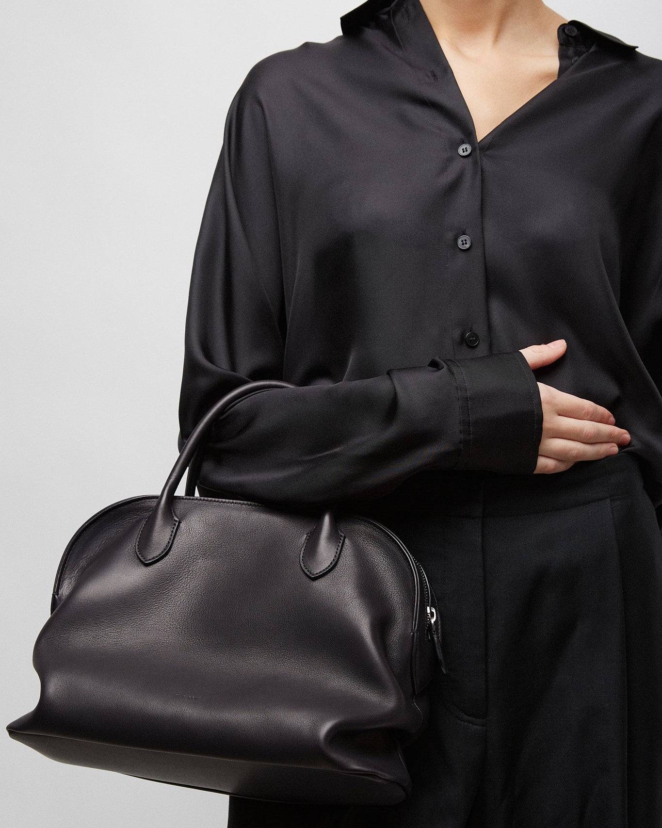 Person wearing a black outfit holding a black handbag against a plain background