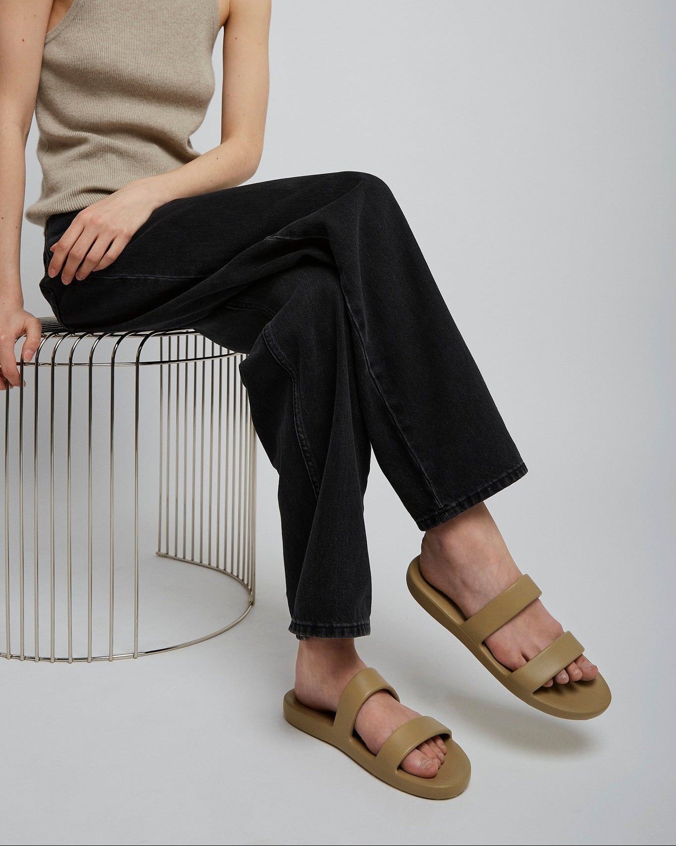 Person wearing beige sandals sitting on a metal stool against a plain background