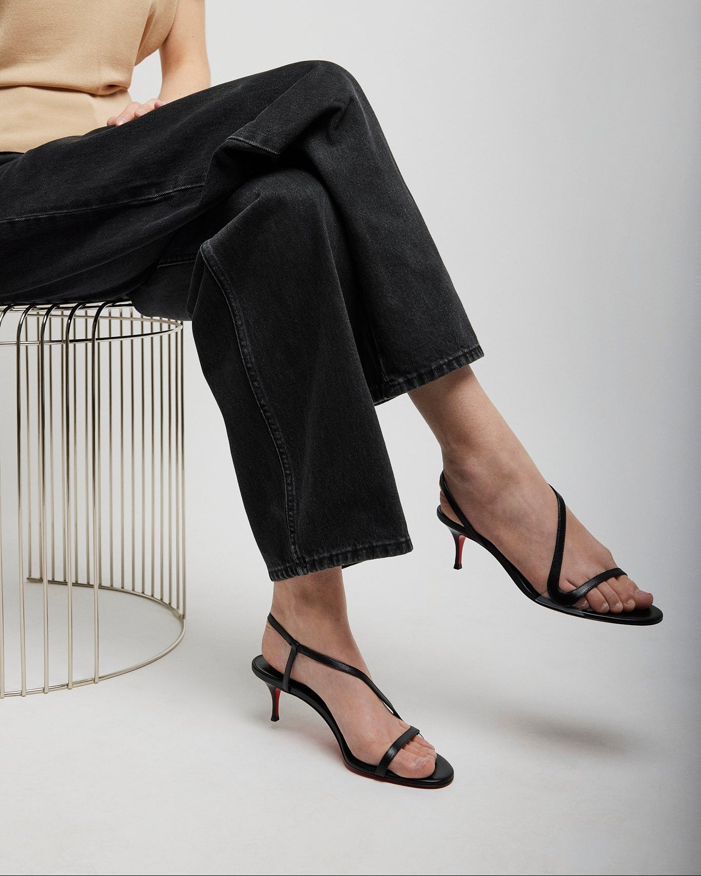 Person wearing black high-heeled sandals sitting on a wire stool against a plain background