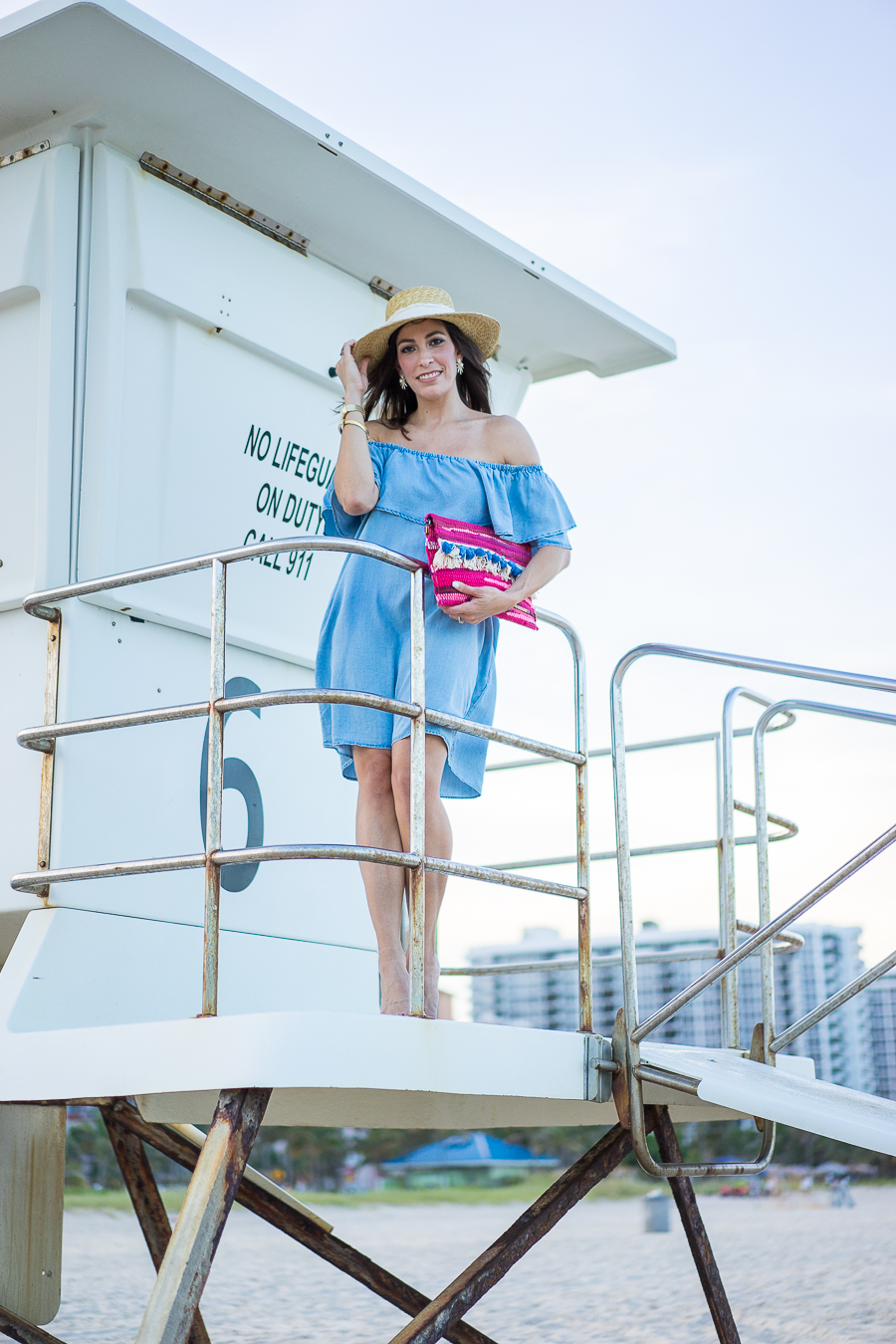 Amanda wearing Soft Chambray Denim Dress and holding her Rag and Bone Laurie straw boater hat and standing at the lifeguard house