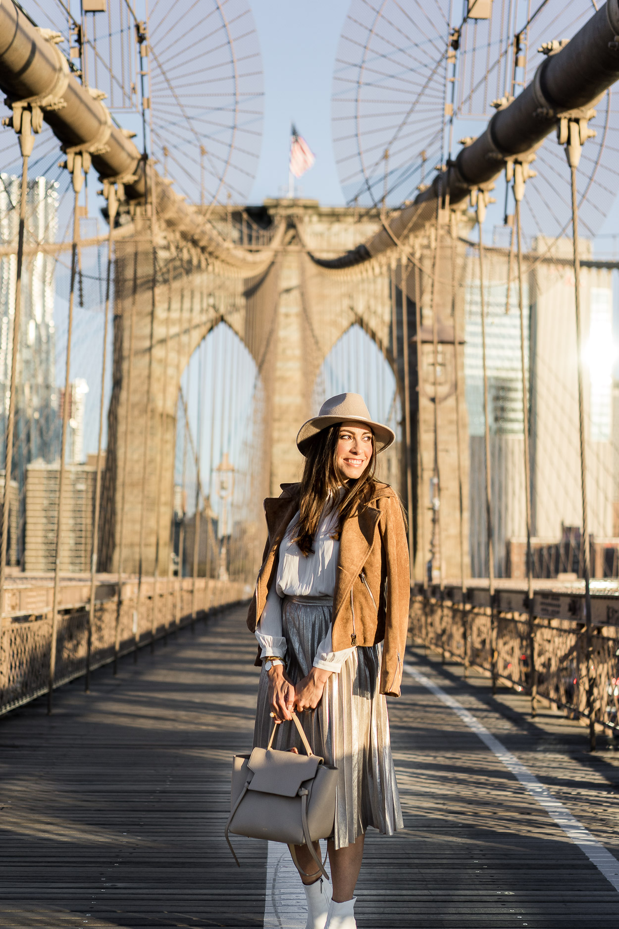 A metallic pleated skirt is a staple piece for Fall styled by Amanda of Best Luxury Designers Handbags with her Celine belt bag, Topshop white booties and a Lulus camel suede jacket at the Brooklyn Bridge